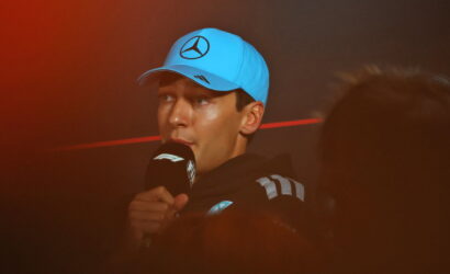 Man in a light-blue cap with a Mercedes logo speaks into a microphone during an interview on a dark stage.
