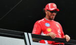 Formula 1 driver in red Ferrari gear with cap and sunglasses standing behind a railing on a balcony at a race event.