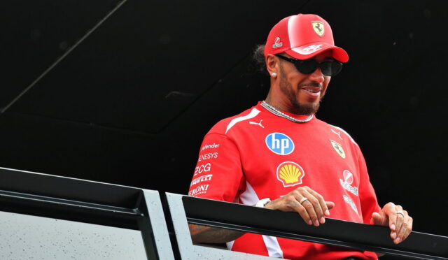 Formula 1 driver in red Ferrari gear with cap and sunglasses standing behind a railing on a balcony at a race event.