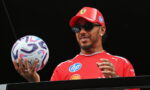 Athlete in a red cap and shirt on a podium, holding a patterned soccer ball.