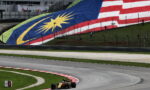 Formula 1 car on a curved race track with a large Malaysian flag mural on the hillside behind it.