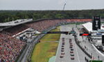 Wide shot of a Formula 1 race start: cars lined up on the starting grid as a packed grandstand watches.