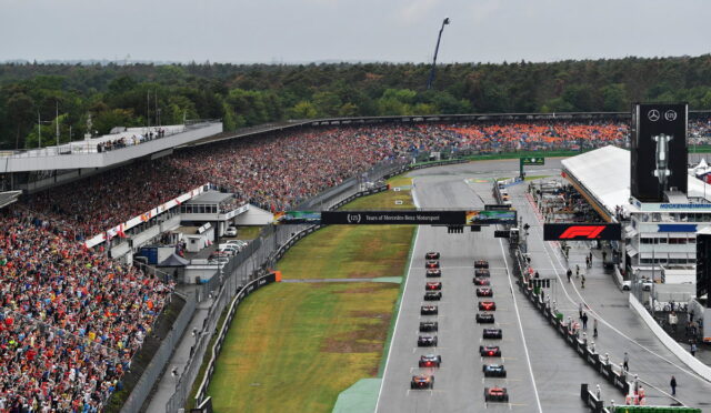 Wide shot of a Formula 1 race start: cars lined up on the starting grid as a packed grandstand watches.