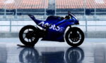 Blue Moto3 racing motorcycle parked on a glossy surface with empty stadium seating in the background