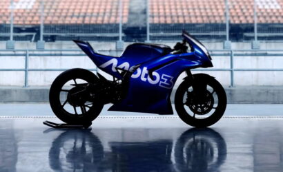 Blue Moto3 racing motorcycle parked on a glossy surface with empty stadium seating in the background