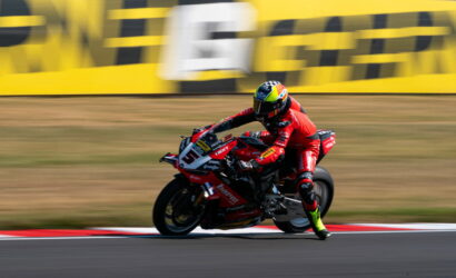Moto racer on a red Ducati motorbike corners at speed on a racetrack, yellow barriers in the background.