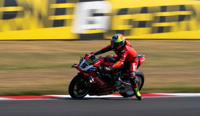 Moto racer on a red Ducati motorbike corners at speed on a racetrack, yellow barriers in the background.