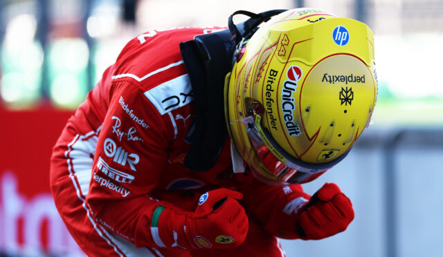Close-up of a race car driver in a red suit and yellow helmet, gripping red racing gloves in a celebratory pose.